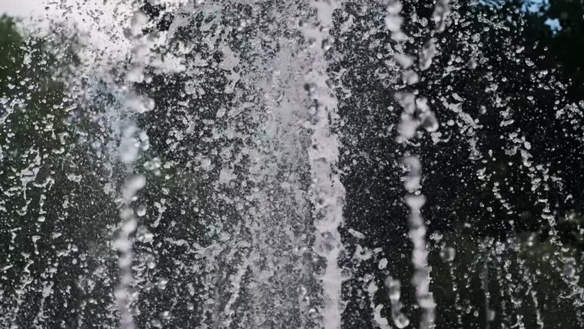 Close-up slow-motion shot of a powerful fountain jet spraying upwards, creating a dense texture of water droplets against a dark, natural background. The scene is energetic and refreshing.