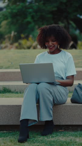 Positive African American woman remote worker types on laptop keyboard in green park. Female freelancer works on notebook computer sitting on parapet. Freelance job. Vertical shot