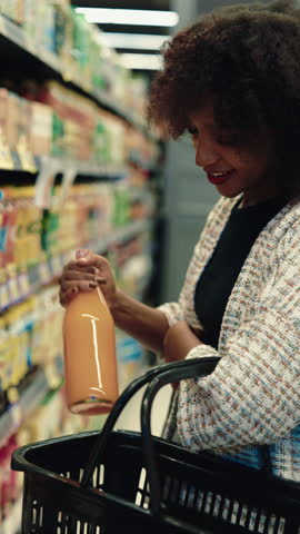 Portrait of positive African American lady placing bottles of juice into basket in store. Black female buys healthy drinks in supermarket. Woman chooses products vertical shot