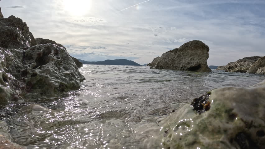 Sunlight reflecting on water surface with rocks. Bright sunlight gleaming on rippling water surface, rocky shoreline and distant island creating serene seascape with peaceful mediterranean atmosphere
