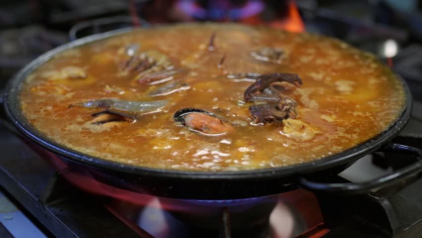 Seafood paella simmering in professional kitchen, vibrant ingredients bubbling within traditional spanish pan over intense gas flame
