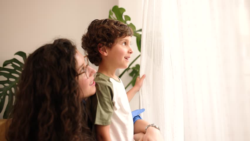 Real time zoom in side view of smiling mother in eyeglasses looking away and conversing with standing son while sitting near window with curtain showing index finger and folding shirt sleeve