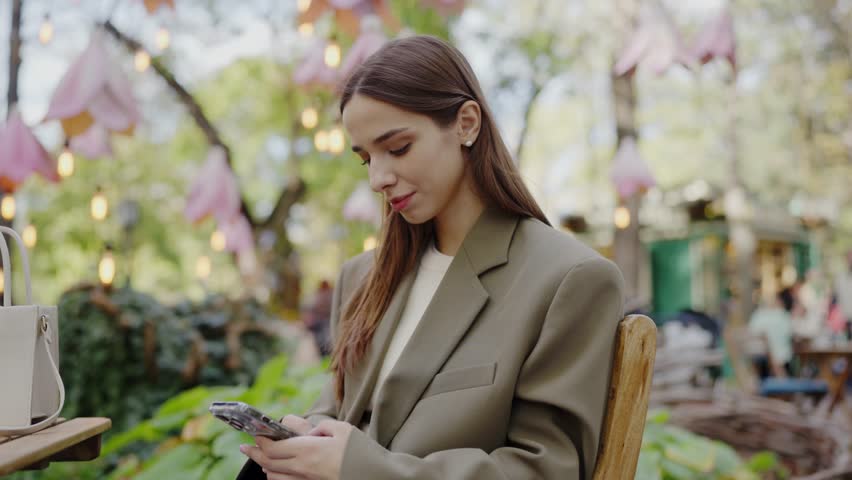 Focused young woman in a stylish beige suit engages with her smartphone at a cozy outdoor cafe surrounded by lush greenery. Young Woman Using Smartphone in Outdoor Cafe Setting