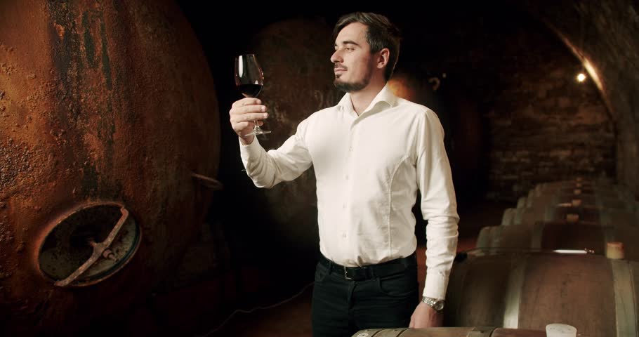 Man tasting wine in a factory or warehouse. Nicely Sommelier checking and drinking a glass of red wine. Successful male winemaker is tasting a flavor and checking fresh red wine in a wine cellar.