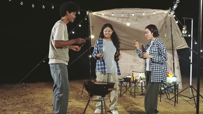 Three people are standing around a grill. One of them is holding a bottle. The grill is on a table