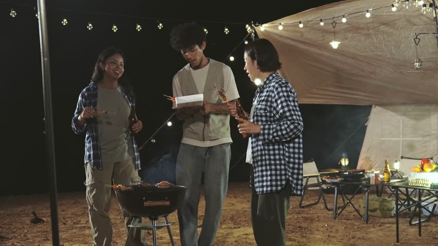Three people are standing around a grill. One of them is holding a bottle. The grill is on a table