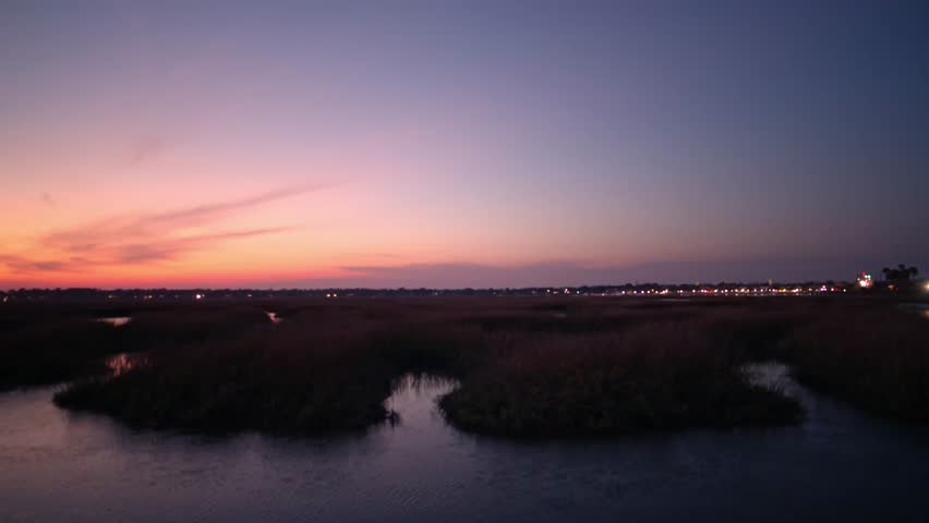 Sunset over wetlands with winding waterways and distant city lights during twilight hours