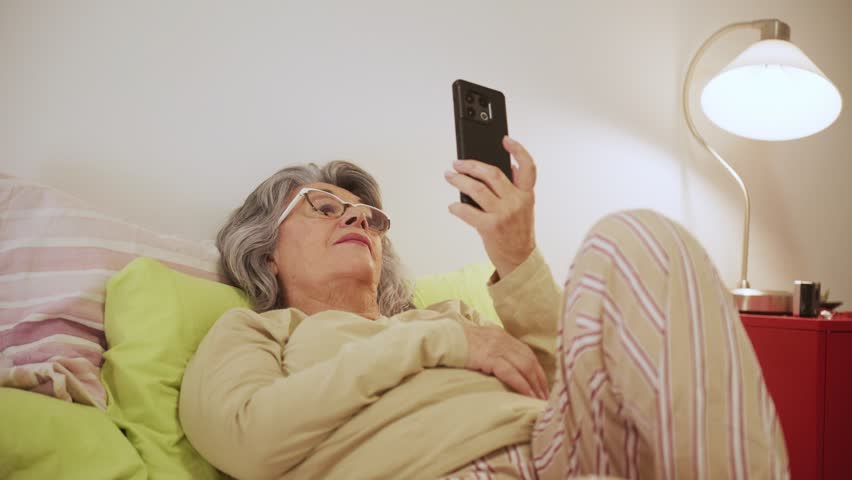 An older woman is comfortably lying in bed, engaging in a video call on her smartphone. The simple setting features soft lighting and a relaxed atmosphere, showcasing a heartfelt connection.