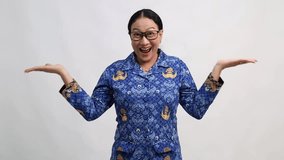 A cheerful Indonesian government worker woman in blue Korpri batik uniform smiles widely and holds her hands out, palms up, as if presenting or weighing options, against a plain white background. - Powered by Shutterstock - Get 15% off with code: PIKWIZARD15