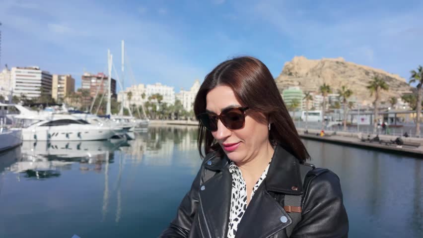 Young woman wearing sunglasses and a black leather jacket using smartphone in alicante port, with luxury yachts and mount benacantil in the background