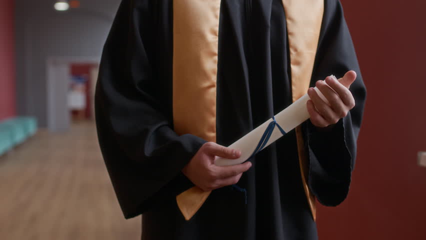 Tilt up portrait of young cheerful male graduating student in academic wear holding diploma smiling in university hall, copy space