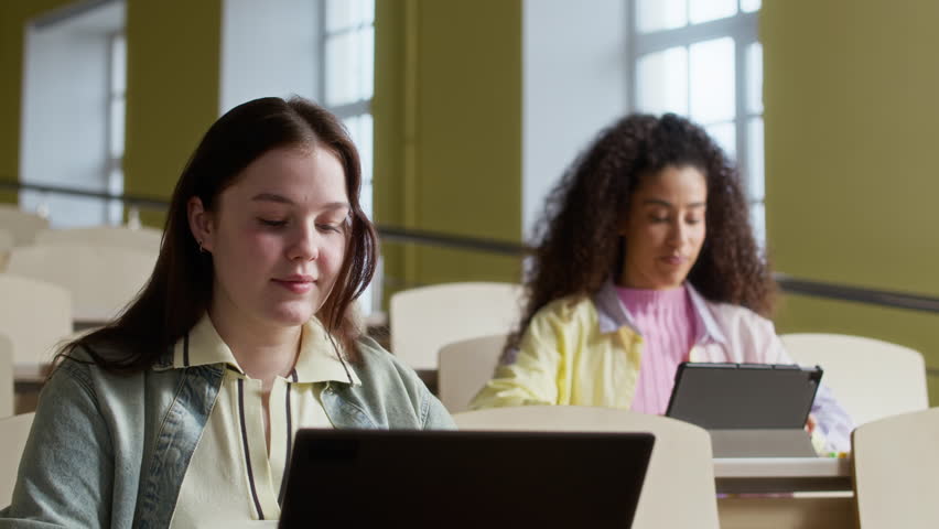 Rack focus of multiethnic female undergraduates sitting at wooden desks, offering each other markers in lecture hall