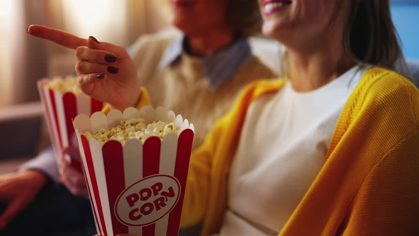 Cheerful young woman and senior mom eating popcorn while watching movie at home