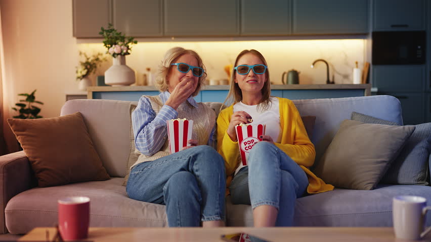Happy young woman and senior mother in 3D glasses eating popcorn, watching movie