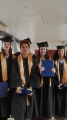 Vertical zoom in portrait of cheerful interethnic graduating students in academic gowns and hats posing for camera with diplomas