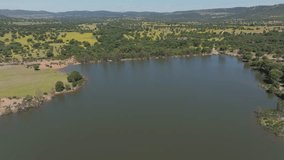 Aerial drone view of green lagoon surrounded by lush vegetation, wildflowers, and oak meadows in Badajoz, Spain. Sunny spring day with blue sky and forested hills on the horizon. - Powered by Shutterstock - Get 15% off with code: PIKWIZARD15
