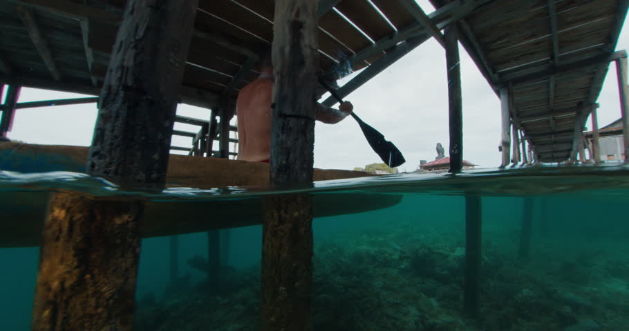 Tattooed caucasian angry man paddles authentic wooden canoe among the stilts in a village of sea gypsy. Angry fit male with tribe tattoos paddle wooden canoe