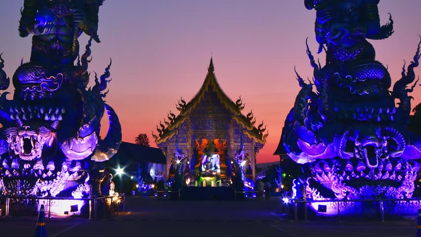 Timelapse zoom out vibrant Blue Temple Wat Rong Suea Ten in Chiang Rai Thailand at sunset with colorful glowing sculptures and dramatic sky