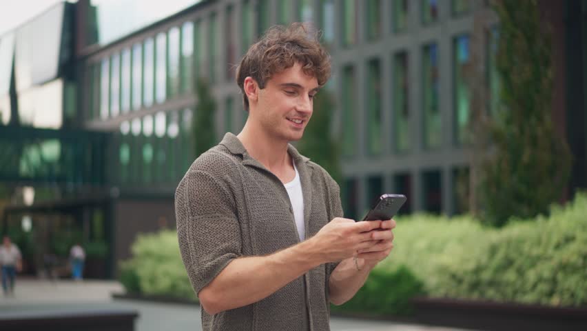 Young happy Caucasian man with curly hair using smartphone while standing outdoors. Male in casual outfit in modern courtyard texting and chatting on mobile phone. Technology and communication.