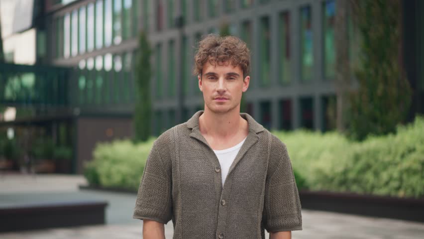 Young happy Caucasian man with curly hair standing confidently outdoors. Male wearing casual outfit looking at camera in modern courtyard. Calm, joyful and composed expression. Portrait. Zooming out.