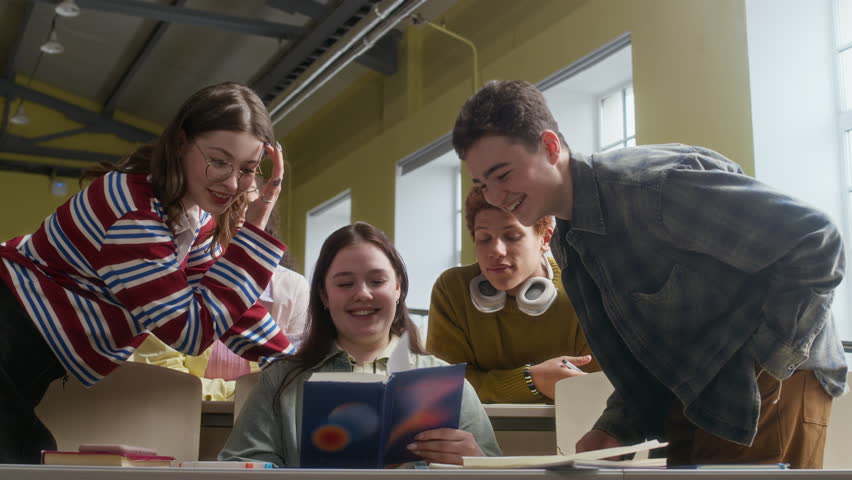 Low angle view of five diverse gen Z peers having fun while reading book in class in college tiered auditorium
