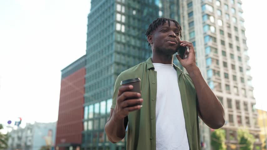 Smiling African American young man talking on mobile phone while holding coffee outdoors. Male in casual outfit standing in city center and chatting on telephone. Enjoying conversation and urban vibes - Powered by Shutterstock - Get 15% off with code: PIKWIZARD15
