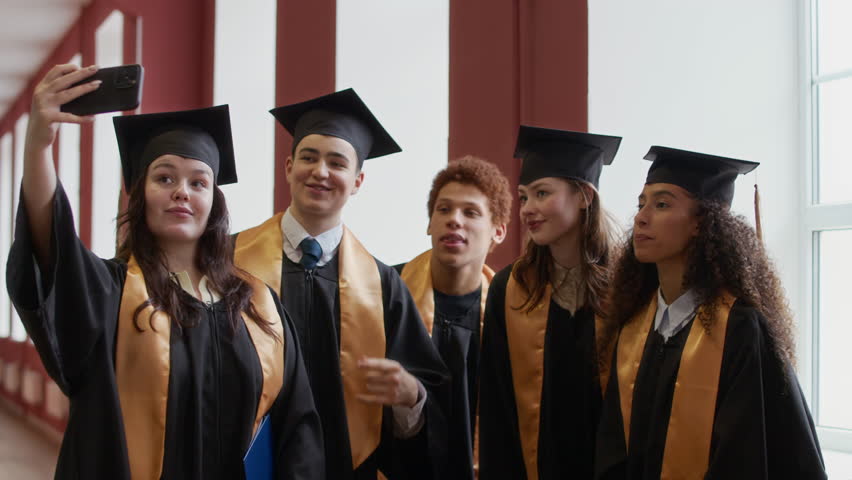Wide shot of five interethnic gen Z graduates wearing black academic gowns and hats taking selfies on smartphone in hall of university