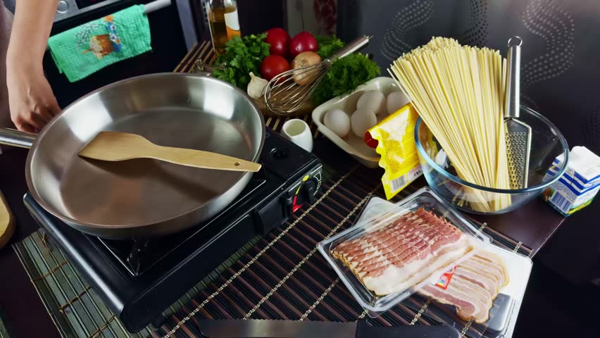 Cooking preparation scene with fresh ingredients on kitchen table. Pasta, eggs, bacon, herbs, tomatoes frying pan ready for making a delicious homemade meal. Pasta carbonara ingredients. Italian food