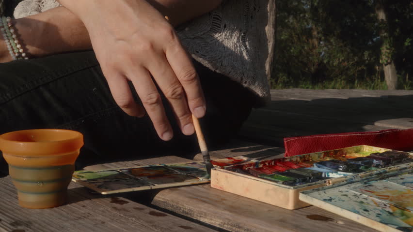 The hand of a female artist with a brush stirs watercolors on a palette on the bank of a river