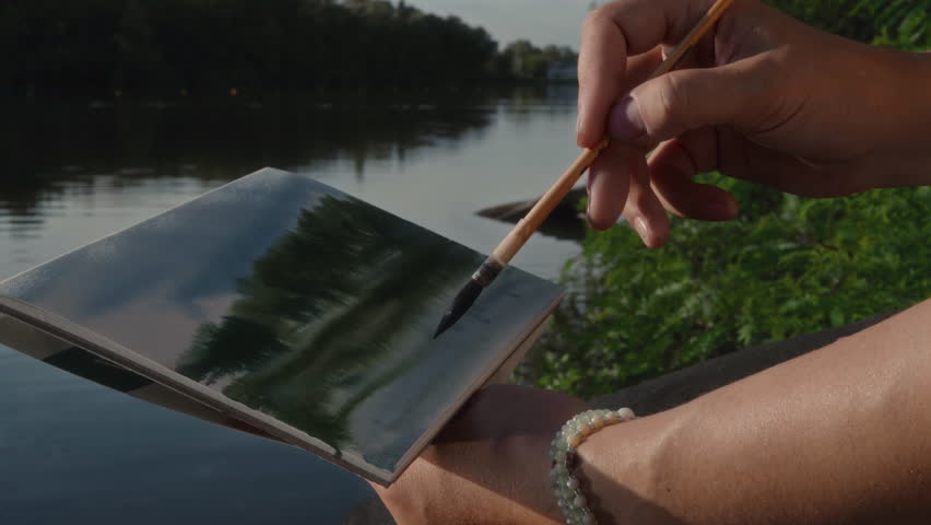 Close-up, female artist painting landscape in watercolor on the river bank