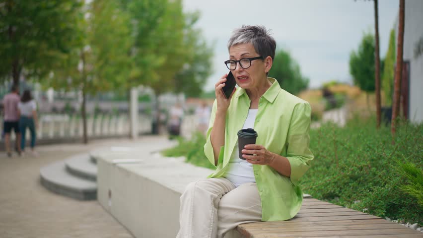 Elderly woman with short gray hair and glasses sitting on bench in park while talking on mobile phone and holding a coffee cup. Caucasian senior female speaking on smartphone and sipping drink.