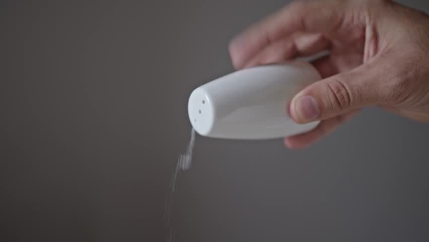 Close up of a hand holding a white salt shaker, sprinkling salt against a plain background - Powered by Shutterstock - Get 15% off with code: PIKWIZARD15