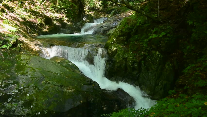 A stunning multi-tiered waterfall called Mie no Taki flows through mossy rocks in Nishizawa Gorge, Yamanashi, Japan. Captured in early autumn, it features clear water and lush green forest.

