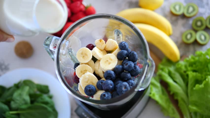 Milk cascading into blender glass filled with colorful banana, blueberry, spinach, strawberry, kiwi, and lettuce ingredients for nutritious smoothie preparation