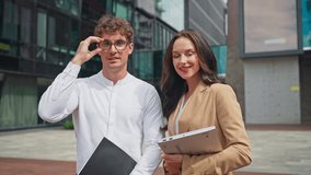 Confident Caucasian man adjusting glasses while holding folder, standing beside elegant woman in beige blazer holding laptop. Smiling and posing together near modern business building. - Powered by Shutterstock - Get 15% off with code: PIKWIZARD15