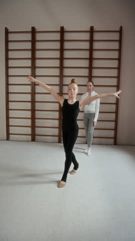 Vertical shot of graceful girl in black tracksuit whirling and keeping balance while practicing new gymnastics exercise, her instructor coming up to her and helping learner