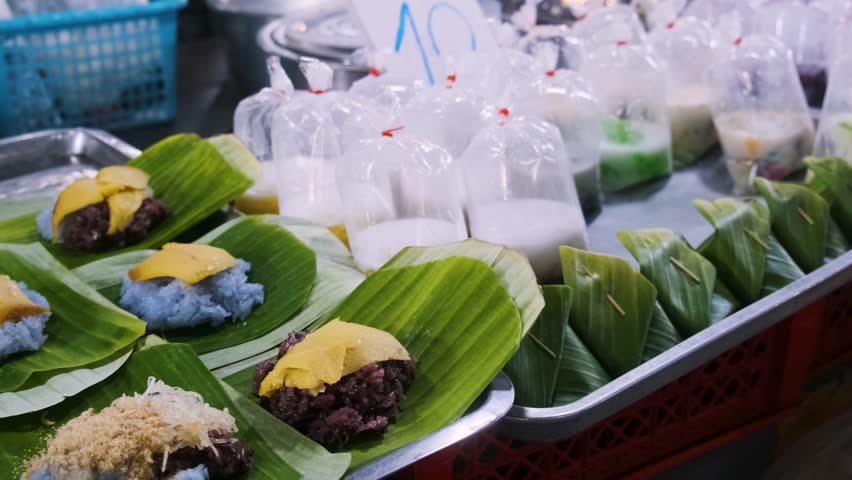 Traditional Thai desserts with coconut milk in plastic bags and sweets wrapped in banana leaves at market stall in night market, Bangkok, Thailand. Thai culinary tradition and take away food in Asia
