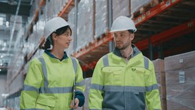 Female warehouse supervisor holding clipboard gesturing upward while explaining task to male coworker. People in high-visibility safety jackets and helmets walking in large industrial storage aisle. - Powered by Shutterstock - Get 15% off with code: PIKWIZARD15