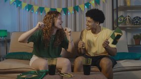 Multiracial couple dancing and cheering during a soccer match at home. The redhead woman and her partner celebrate with energy and rhythm, wearing Brazil colors in a festive and joyful atmosphere. - Powered by Shutterstock - Get 15% off with code: PIKWIZARD15