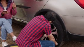 Stranded couple working together during roadside emergency, man replacing flat tire while woman sheltering under umbrella and using mobile phone for potential assistance - Powered by Shutterstock - Get 15% off with code: PIKWIZARD15