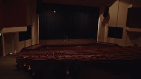 Wide shot showing rows of empty red cushioned seats in a dark theater facing a wooden stage with black curtains and a stool - Powered by Shutterstock - Get 15% off with code: PIKWIZARD15