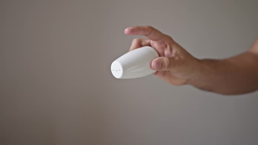 Close up view of a man’s hand sprinkling salt from a white ceramic shaker. Salt falls in a steady stream with a neutral, plain background, highlighting the motion and granules - Powered by Shutterstock - Get 15% off with code: PIKWIZARD15