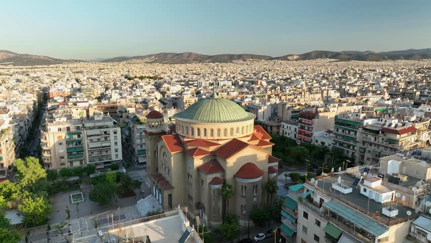 4K aerial view circling the Church of Saint Panteleimon in Athens at sunset. Iconic Greek Orthodox architecture featuring a green dome and red-tiled roof, framed by the glowing urban landscape.