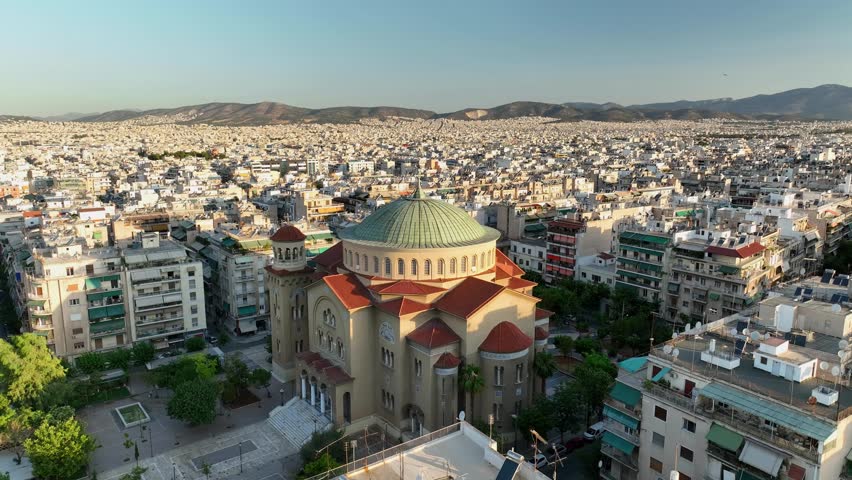 UHD 4K drone shot of Church of Saint Panteleimon of Acharnai in Athens, Greece. Greek Orthodox cathedral with green dome and red roof, surrounded by dense urban cityscape at golden hour.