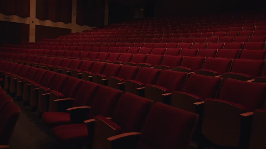 Wide view of tiered rows of empty red theater seats inside a dimly lit auditorium. Slow motion view