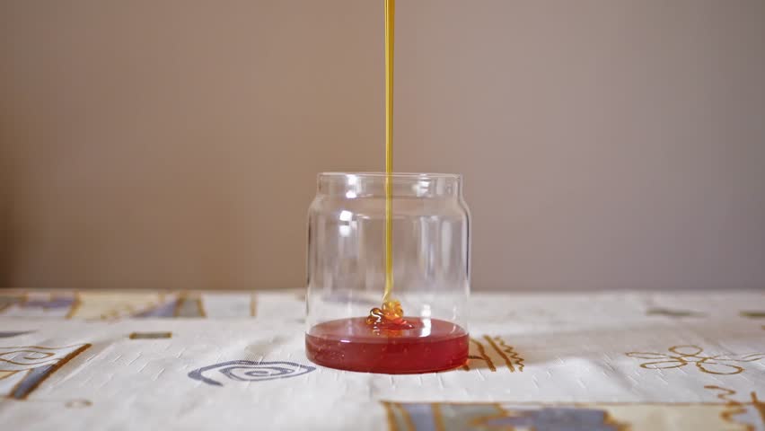 Thick golden honey pours in slow motion from above into an empty glass jar, accumulating in a smooth pool. Close up view
