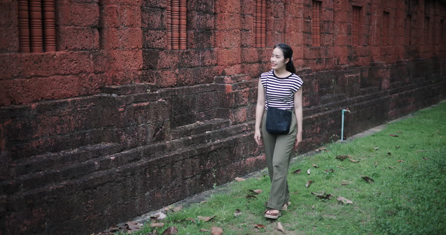Handheld high angle view shot, Relaxed young woman wearing a striped shirt and green pants walking around, admiring the natural scenery and ancient brick structures, Enjoy holiday travel.