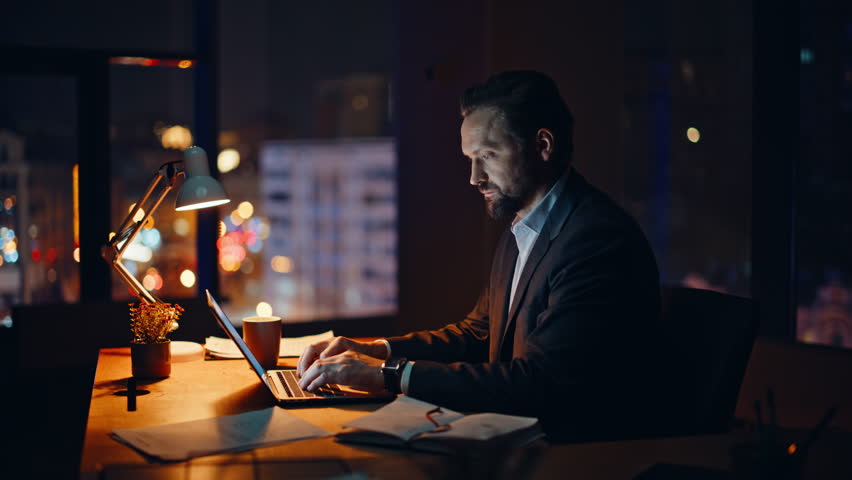 Overworked man typing laptop checking papers at evening workspace closeup. Serious businessman working late looking computer in office. Smart lawyer thinking decision analyzing reports alone at night 