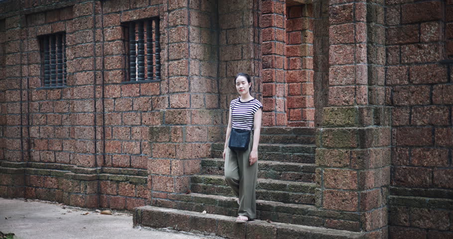 Handheld shot, Relaxed young woman wearing a striped shirt and green pants walking around, admiring the natural scenery and ancient brick structures, Enjoy holiday travel.