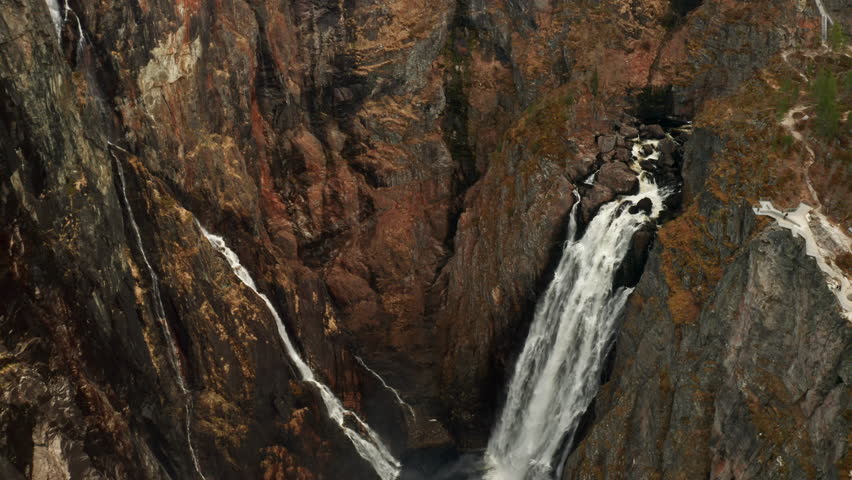 Powerful Voringsfossen waterfall cascades down rugged cliff face into dark pool below. Canyon walls display mix of earthy tones with streaks of water